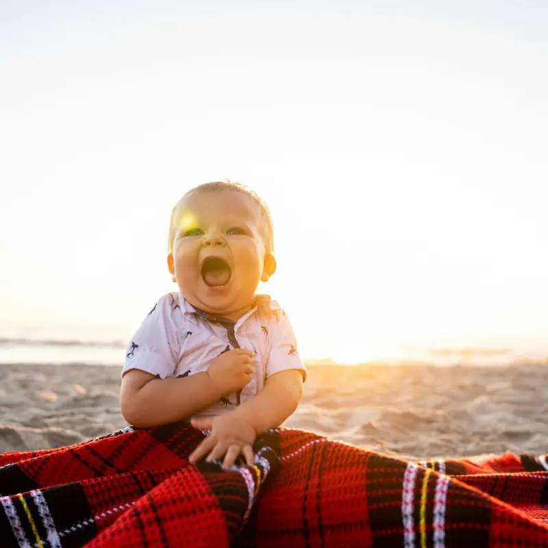 bébé riant sur une plage en été, assis sur un tissu rouge au coucher du soleil