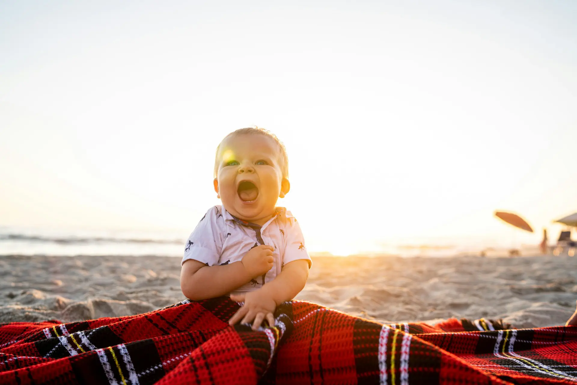 bébé riant sur une plage en été, assis sur un tissu rouge au coucher du soleil